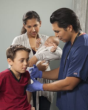 Healthcare provider giving boy an injection. Mother holding baby in background.